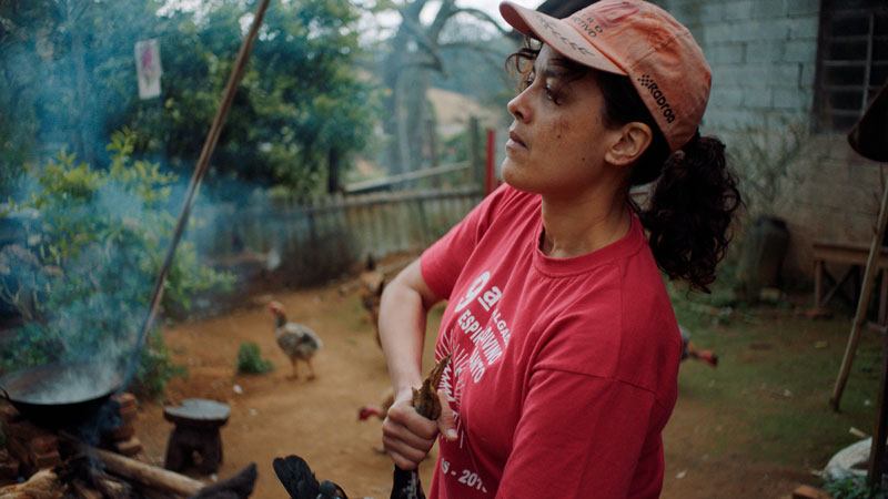 Uma mulher negra cabelos cacheados vestindo boné e camiseta vermelhos segura uma galinha pelos pés. A imagem é uma cena do filme Carvão, de Carolina Markowicz.